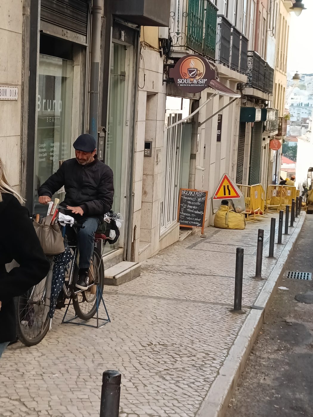 A man on a bicycle on a cobbled pavement lined with bollards, outside a row of shops