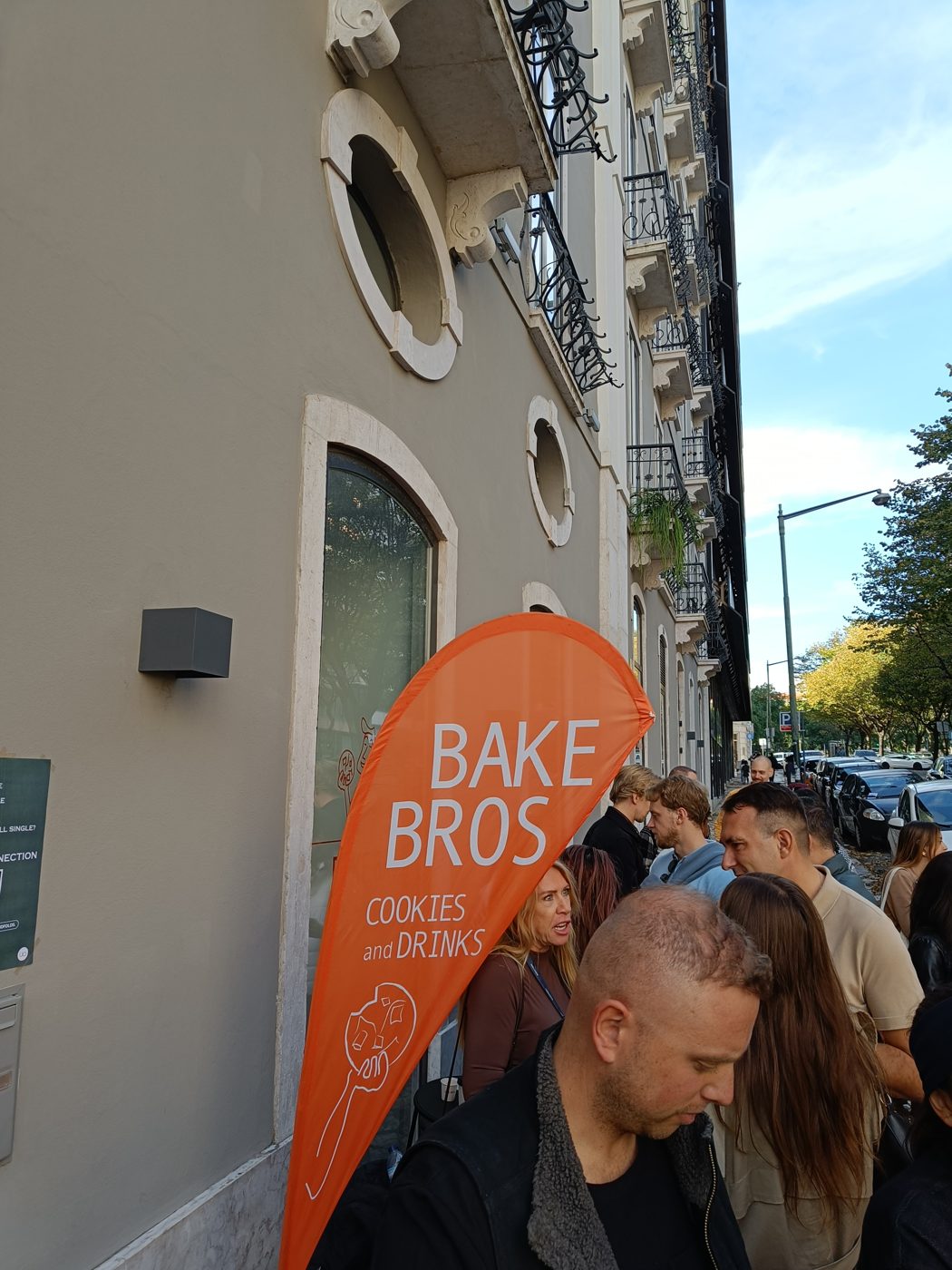 People crowding a pavement beside an orange bakery sign
