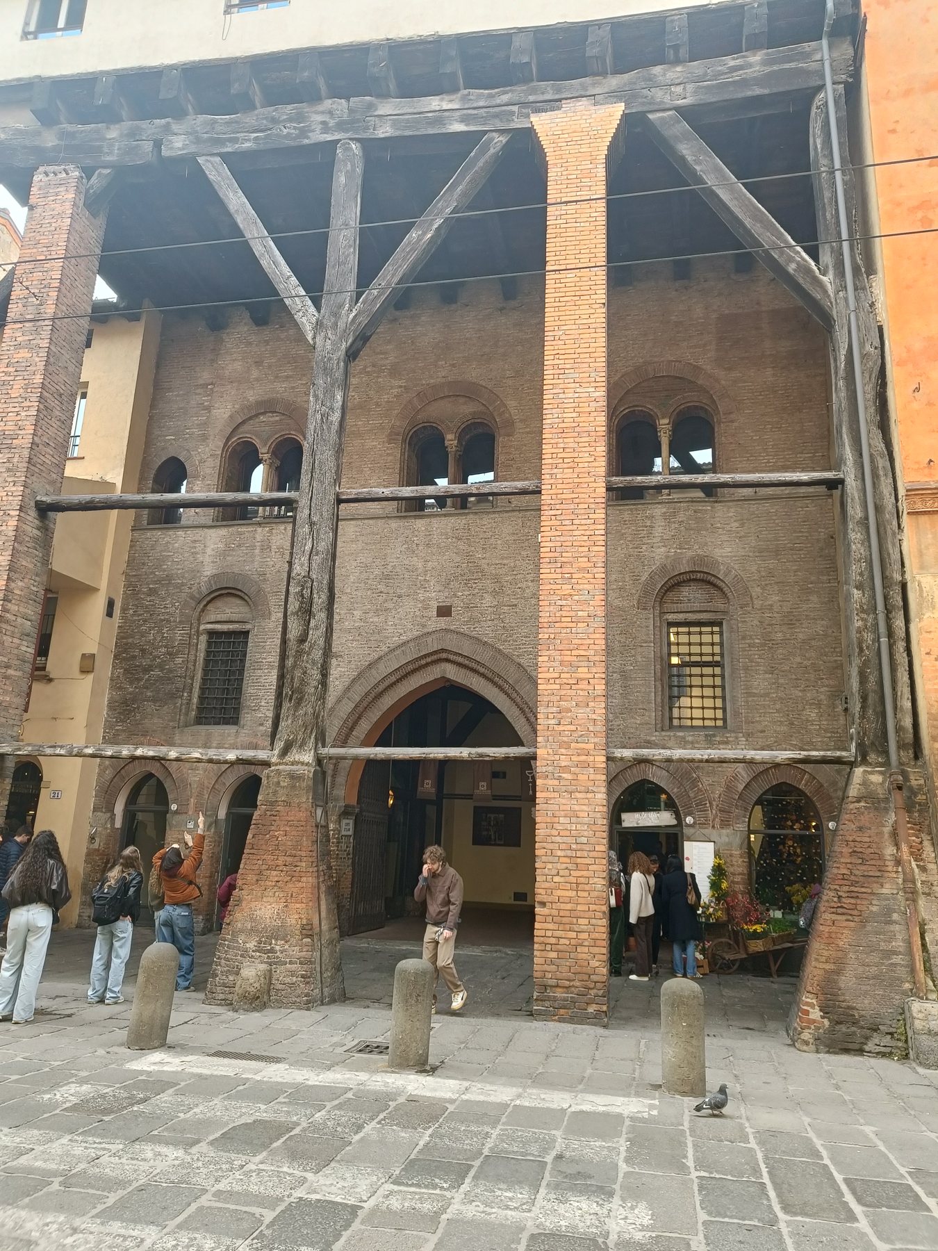 A medieval building front propped up with timber beams, people walking underneath