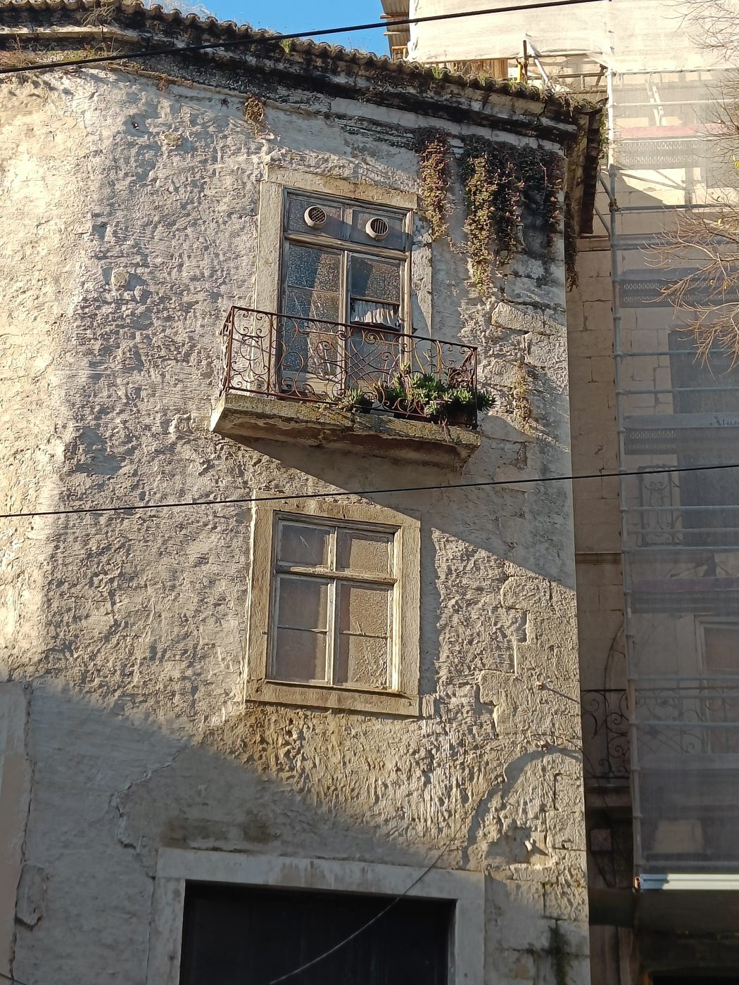 A weathered pale stone house front with shuttered windows and a small balcony, scaffolding beside it