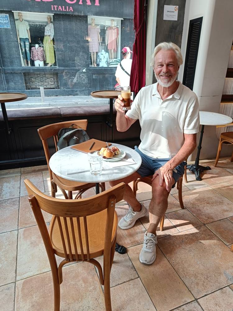 Hans at a café table in Athens, raising a coffee