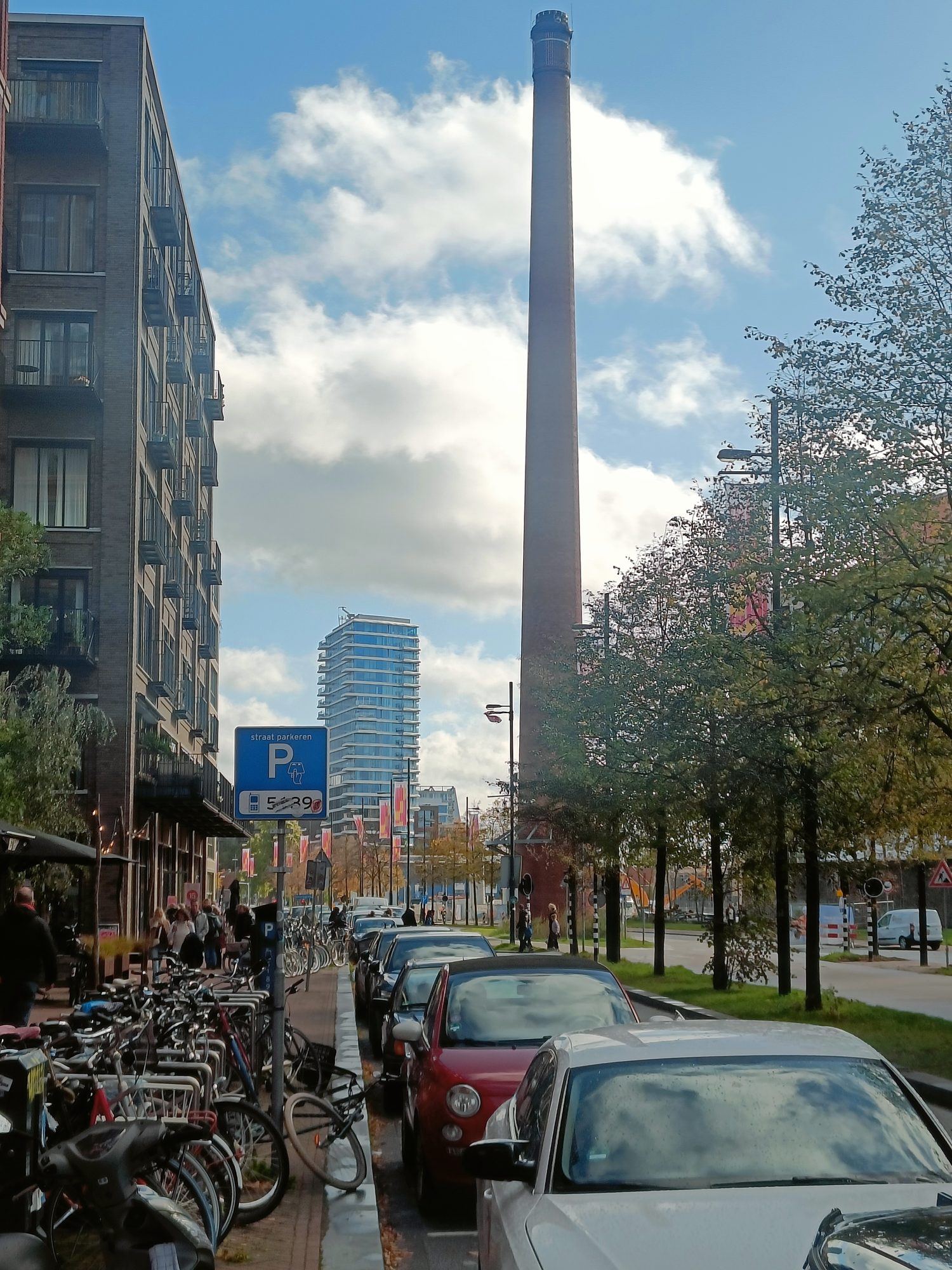 An old factory chimney between new apartment blocks, with bikes and parked cars along the street