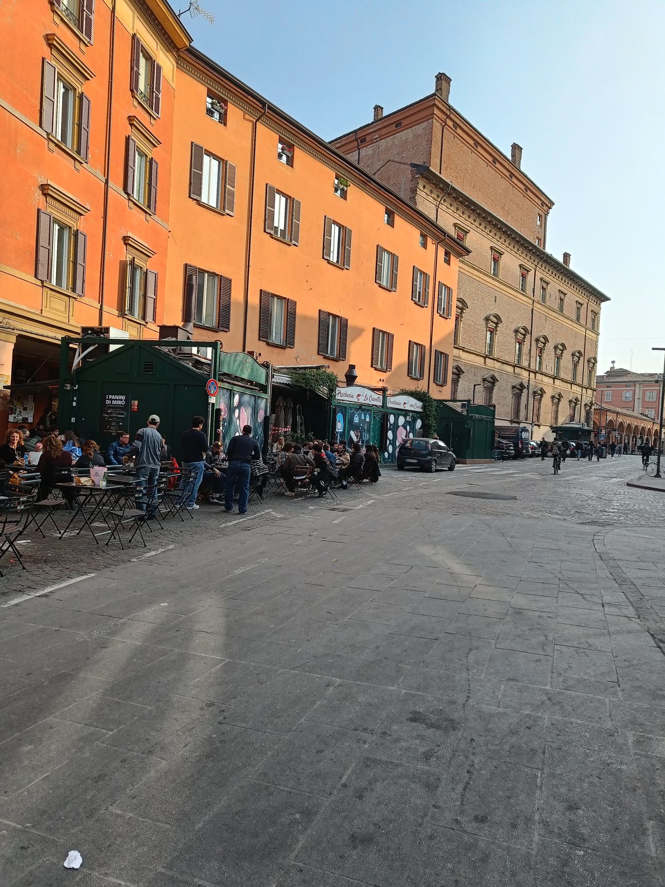 An orange row of buildings over a wide street, with people at kiosk tables in the sun