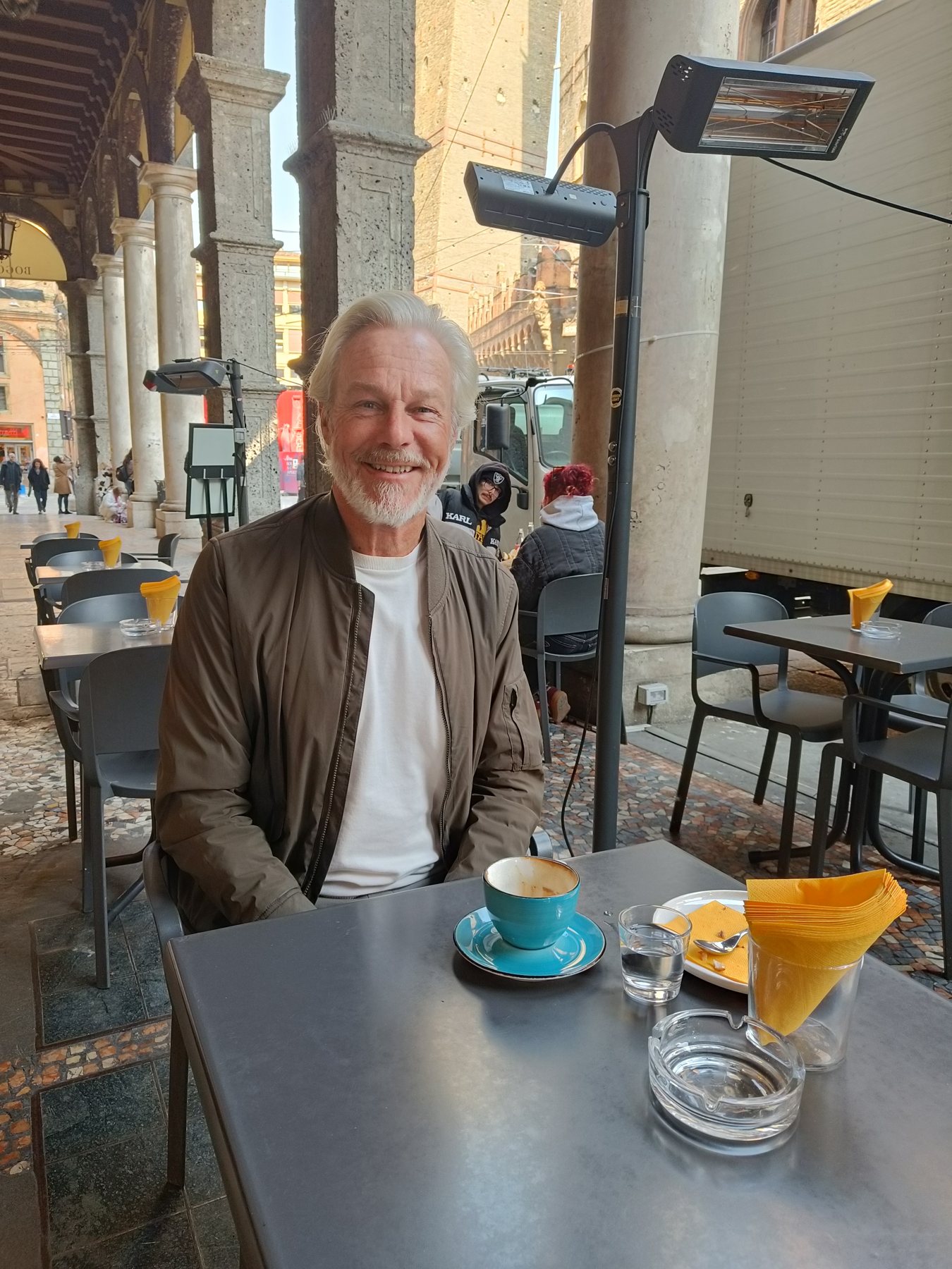 Hans at a café table under the porticoes with a coffee, smiling